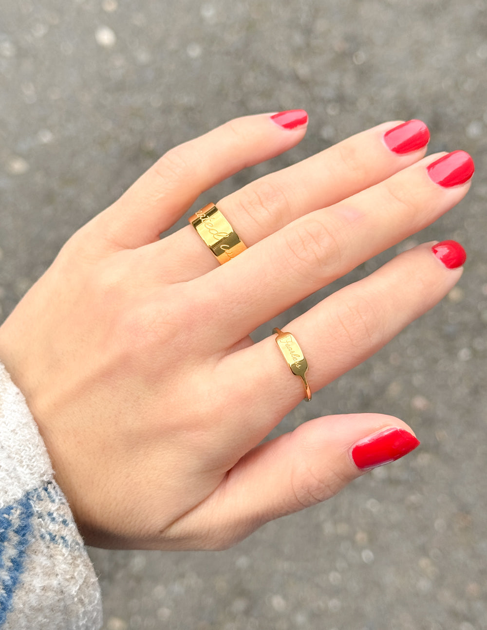 Hand with red nail polish wearing two gold rings on a blurred background