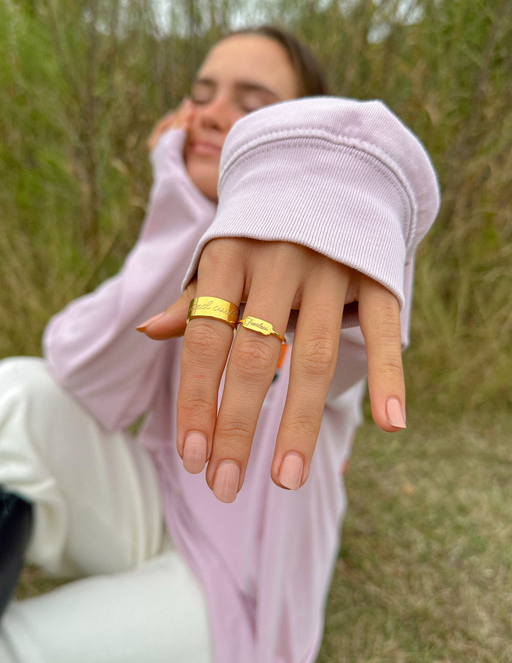 Person wearing a light pink sweatshirt and white pants, showing off gold rings outdoors.