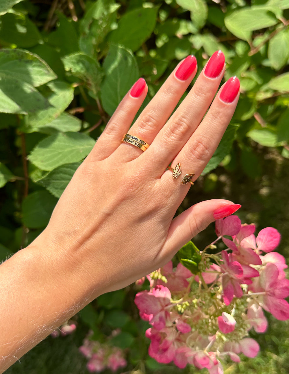 Hand with pink nails and rings in front of green foliage and pink flowers