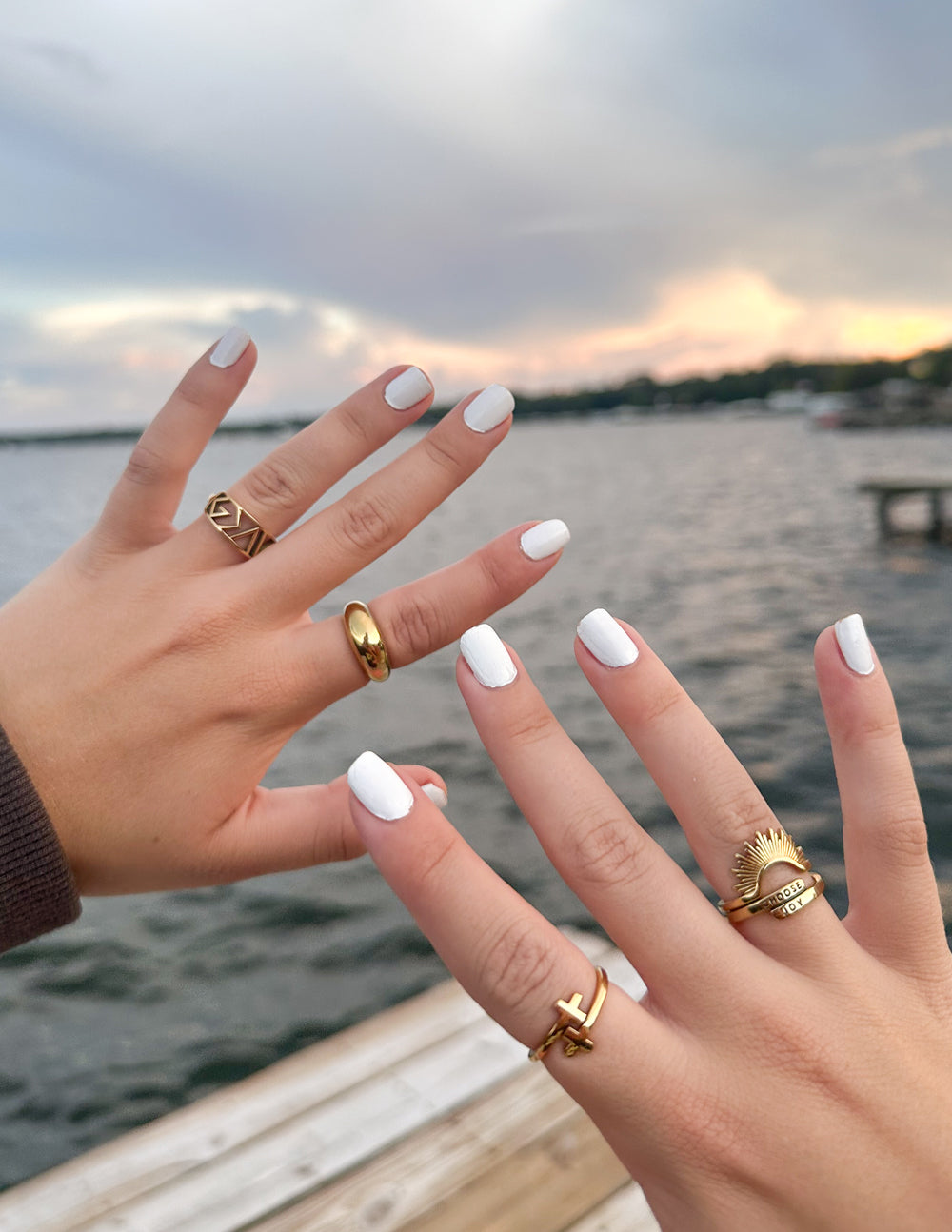 Two hands with gold rings and white nail polish against a sunset over water background