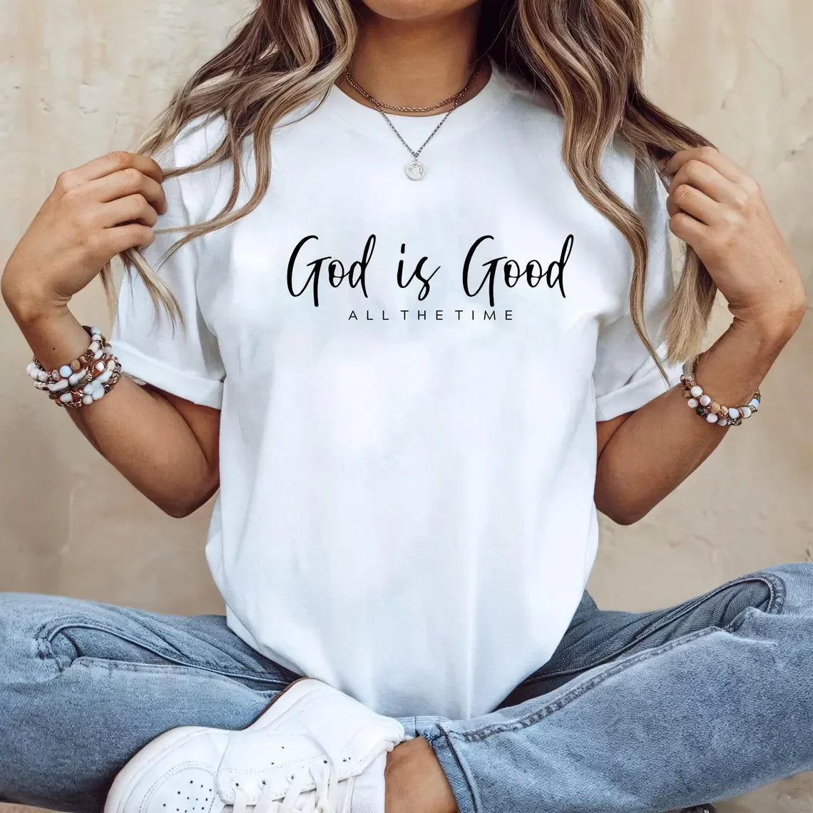 Person wearing a white t-shirt with 'God is Good ALL THE TIME' text, sitting against a neutral background.
