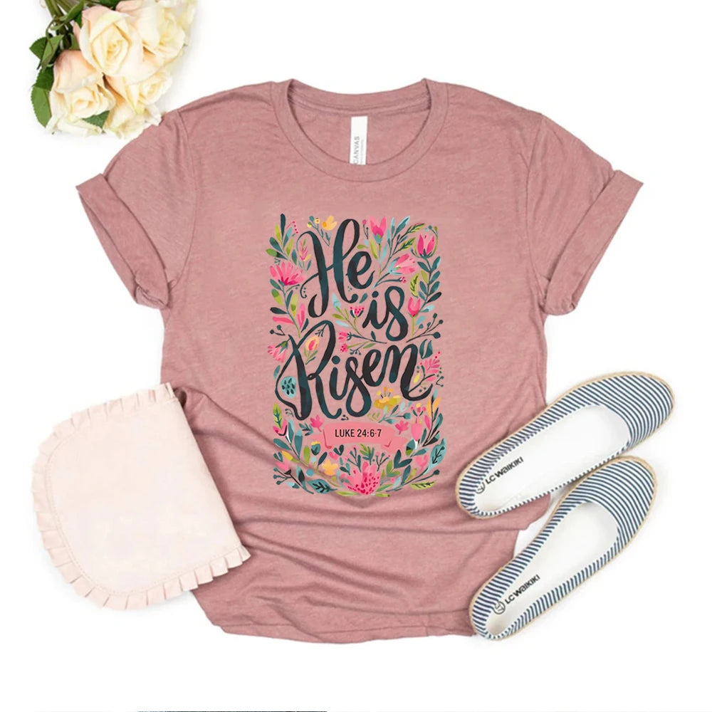 Pink t-shirt with 'He is Risen' design, white slippers, and flowers on a white background