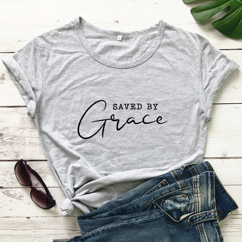 Gray t-shirt with 'Saved by Grace' text on a wooden surface with jeans and sunglasses.
