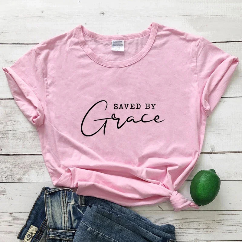 Pink t-shirt with 'Saved by Grace' text on a wooden surface with jeans and a lime.