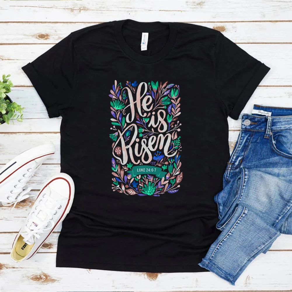 Black t-shirt with 'He is Risen' design on a wooden surface with jeans and sneakers.