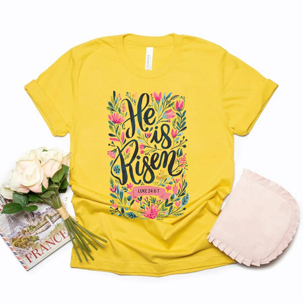 Yellow t-shirt with floral 'He is Risen' design, next to a book and flowers on a white background