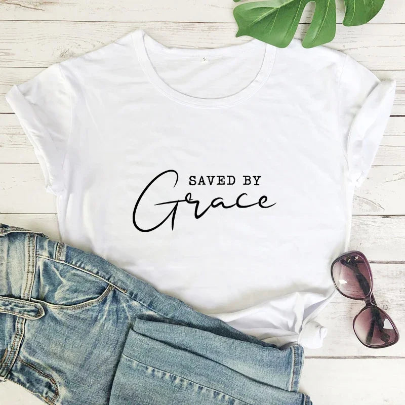 White t-shirt with 'Saved by Grace' text, denim jeans, and sunglasses on a wooden surface.