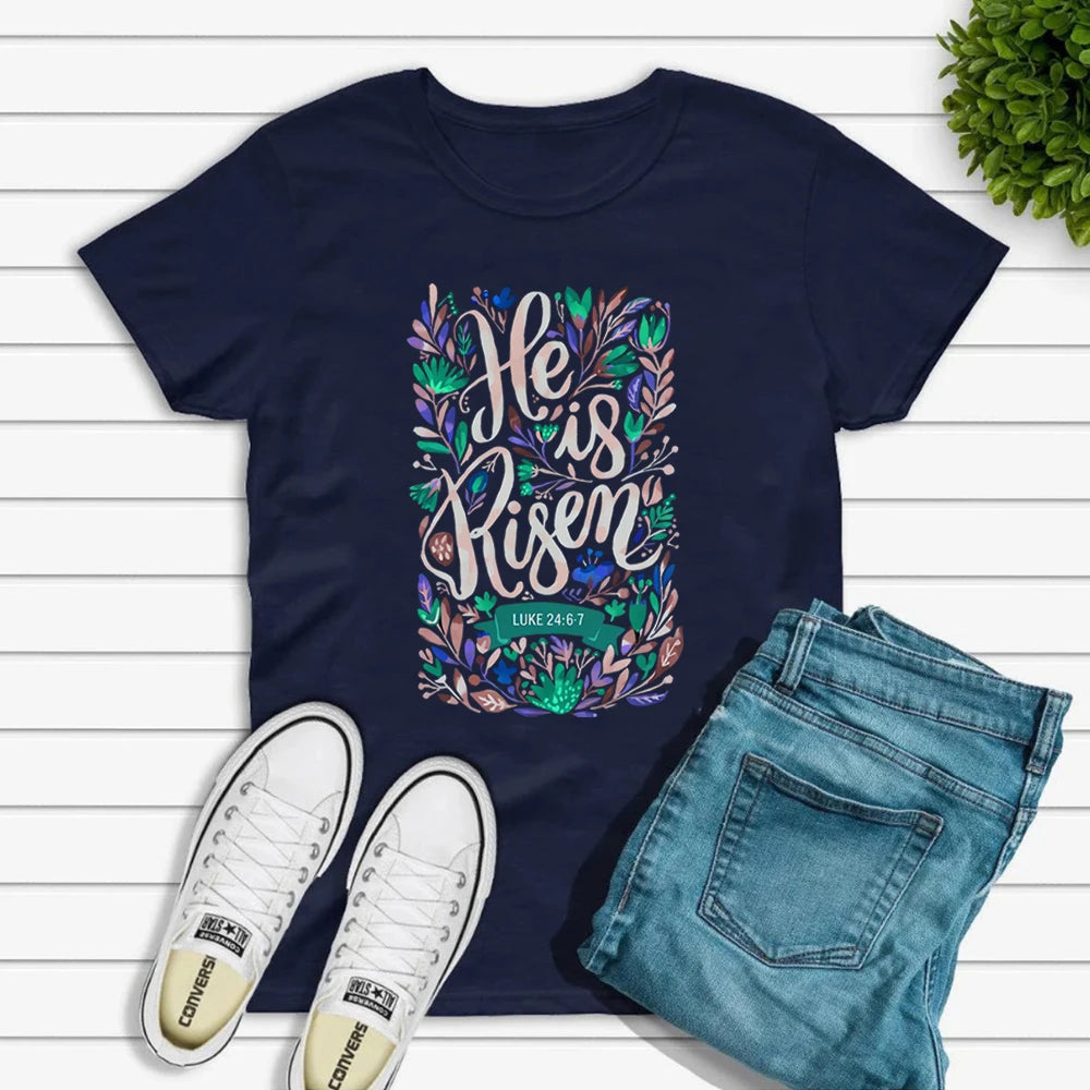 Navy blue t-shirt with 'He is Risen' design, white sneakers, and blue jeans on a wooden surface.
